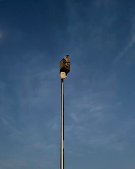 A tall metal pole extends into the sky, topped with a security camera. The background features a clear blue sky with subtle cloud formations.