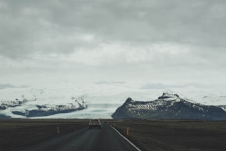 A car is driving on a long, empty road with a dramatic backdrop of snow-capped mountains and a vast glacier under a cloudy, overcast sky.