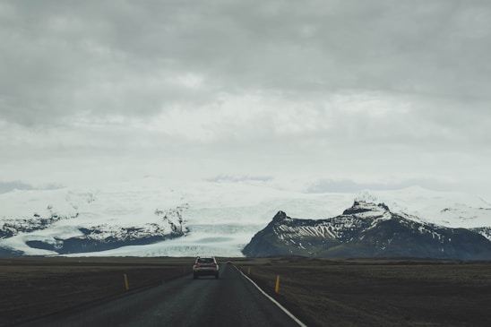 A car is driving on a long, empty road with a dramatic backdrop of snow-capped mountains and a vast glacier under a cloudy, overcast sky.
