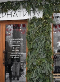 A storefront decorated with lush green branches and string lights. The glass door displays text in Cyrillic, advertising a coffee spot with operating hours and social media information. The surrounding area has a wintry ambiance with snow visible.