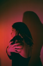 A person with long hair is embracing themselves with their arms wrapped around their shoulders. The light casts a dramatic shadow on the wall, creating an intriguing and introspective visual. The lighting features a blend of pink and red hues, adding a warm and intimate atmosphere.