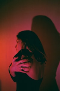 A person with long hair is embracing themselves with their arms wrapped around their shoulders. The light casts a dramatic shadow on the wall, creating an intriguing and introspective visual. The lighting features a blend of pink and red hues, adding a warm and intimate atmosphere.