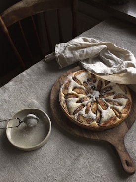 A rustic apple pie dusted with powdered sugar sits on a round wooden board. A light beige linen cloth is casually placed beside it. Nearby, a small ceramic bowl holds a tea strainer filled with powdered sugar. The setting is on a textured, neutral-colored tablecloth, accompanied by a wooden chair in the background.