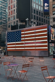 An urban scene featuring a large American flag display on a digital screen, set against the backdrop of tall city buildings. The area is sparsely populated with red metal chairs and tables, suggesting an outdoor seating area. The Taco Bell logo is visible on a building in the background.