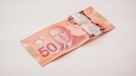 A Canadian 50-dollar bill is resting on a light-colored surface. The note is primarily red and features intricate designs, including a portrait of a historical figure.