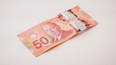 A Canadian 50-dollar bill is resting on a light-colored surface. The note is primarily red and features intricate designs, including a portrait of a historical figure.
