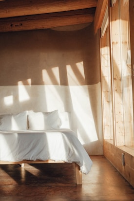 A cozy bedroom features a wooden bed with white bedding and pillows, illuminated by natural sunlight streaming through large windows. The room has a minimalist design with wooden beams on the ceiling and earthy tones.
