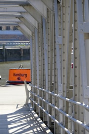 A metal walkway with riveted beams extends into the distance, creating a pattern of light and shadow on the ground. A bright orange buoy with the word 'Hamburg' is visible in the background, partially obstructed by the structure.