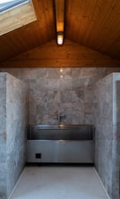 A polished metal sink is set against a backdrop of gray marble tiles in a room with a wooden ceiling. The ceiling features a central light fixture that gives a warm, cozy ambiance. The floor is made of small hexagonal white tiles.
