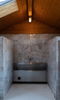 A polished metal sink is set against a backdrop of gray marble tiles in a room with a wooden ceiling. The ceiling features a central light fixture that gives a warm, cozy ambiance. The floor is made of small hexagonal white tiles.