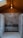 A polished metal sink is set against a backdrop of gray marble tiles in a room with a wooden ceiling. The ceiling features a central light fixture that gives a warm, cozy ambiance. The floor is made of small hexagonal white tiles.