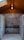 A polished metal sink is set against a backdrop of gray marble tiles in a room with a wooden ceiling. The ceiling features a central light fixture that gives a warm, cozy ambiance. The floor is made of small hexagonal white tiles.