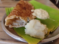 A crispy brown dosa placed on a banana leaf with a serving of white chutney and mashed potatoes on a metal plate.
