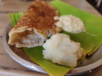 A crispy brown dosa placed on a banana leaf with a serving of white chutney and mashed potatoes on a metal plate.