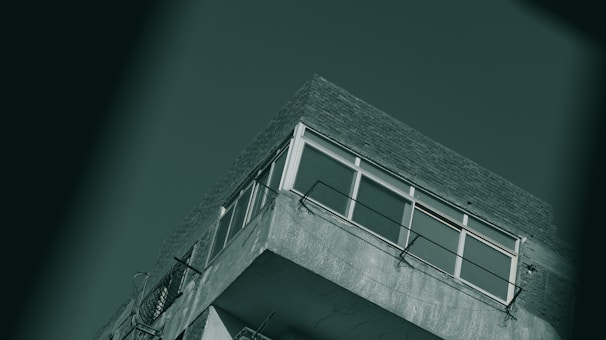 A section of a concrete building featuring a balcony with metal railings, windows that appear to be office-like, and a flat roof with a brick facade against a solid background.