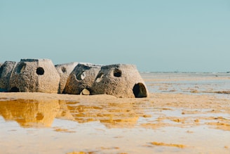 Concrete structures with circular openings rest on a sandy beach, partially submerged by shallow water. The scene is set under a clear blue sky, creating a peaceful and serene coastal environment.