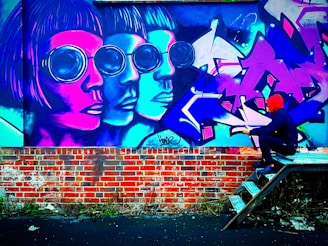 Vivid graffiti art adorns a brick wall, featuring three overlapping and stylized human faces with large, round sunglasses in shades of pink, blue, and purple. A person in a red hoodie and dark clothing is sitting on metal steps facing the mural, appearing to engage with it. The scene is lively with intense colors.