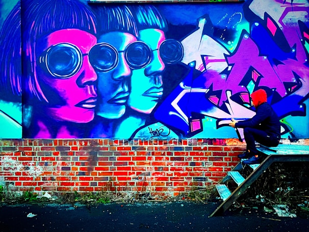 Vivid graffiti art adorns a brick wall, featuring three overlapping and stylized human faces with large, round sunglasses in shades of pink, blue, and purple. A person in a red hoodie and dark clothing is sitting on metal steps facing the mural, appearing to engage with it. The scene is lively with intense colors.
