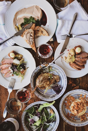 An arrangement of plates with various gourmet dishes on a wooden table. There are slices of bread with p&acirc;t&eacute;, a dish of duck confit, a plate with sliced meat and salad, and a dish with dessert topped with almonds. The scene includes glasses of red wine and is set with white napkins and silverware.