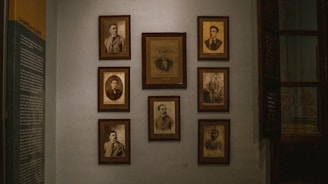 A dimly lit wall decorated with several framed vintage sepia-toned portraits. The portraits are arranged in a neat grid and feature individuals in formal attire, typical of the early to mid-20th century. On the left side of the wall, there is a text panel providing information, likely related to the people in the photographs.