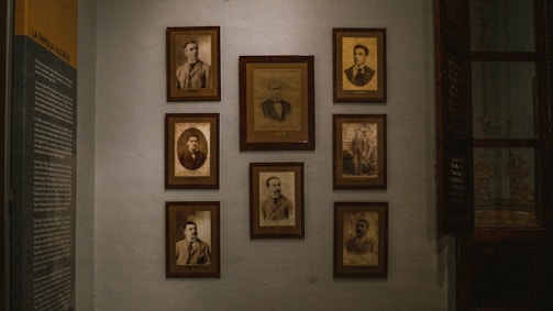 A dimly lit wall decorated with several framed vintage sepia-toned portraits. The portraits are arranged in a neat grid and feature individuals in formal attire, typical of the early to mid-20th century. On the left side of the wall, there is a text panel providing information, likely related to the people in the photographs.
