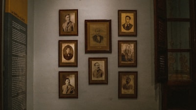 A dimly lit wall decorated with several framed vintage sepia-toned portraits. The portraits are arranged in a neat grid and feature individuals in formal attire, typical of the early to mid-20th century. On the left side of the wall, there is a text panel providing information, likely related to the people in the photographs.