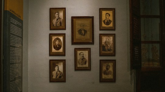 A dimly lit wall decorated with several framed vintage sepia-toned portraits. The portraits are arranged in a neat grid and feature individuals in formal attire, typical of the early to mid-20th century. On the left side of the wall, there is a text panel providing information, likely related to the people in the photographs.