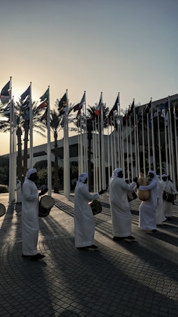 A group of people dressed in traditional white garments performs with drums and other percussion instruments. The individuals are arranged in a line, creating rhythmic patterns as the sunlight casts long shadows on the cobblestone surface. Numerous national flags are raised on poles in the background, with palm trees silhouetted against the setting sun.