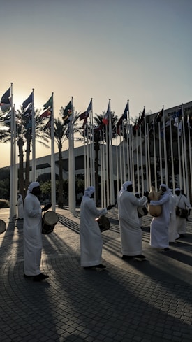 A group of people dressed in traditional white garments performs with drums and other percussion instruments. The individuals are arranged in a line, creating rhythmic patterns as the sunlight casts long shadows on the cobblestone surface. Numerous national flags are raised on poles in the background, with palm trees silhouetted against the setting sun.