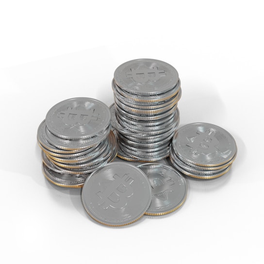 A collection of silver-colored coins stacked in piles, each coin bearing a Bitcoin symbol. The coins are arranged on a white surface, creating a reflective and metallic effect.