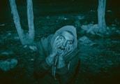 A person wearing a hooded jacket displays a frightened expression, pressing their hands against their face. The eyes appear unnaturally rolled back. The surroundings are dimly lit, with trees and rocks in the background, giving a creepy or horror-like atmosphere.