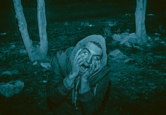 A person wearing a hooded jacket displays a frightened expression, pressing their hands against their face. The eyes appear unnaturally rolled back. The surroundings are dimly lit, with trees and rocks in the background, giving a creepy or horror-like atmosphere.