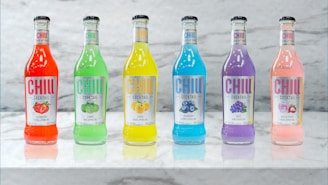 Six colorful cocktail bottles are arranged in a line on a marble surface. Each bottle is filled with a different brightly colored drink: red, green, yellow, blue, purple, and pink. The labels on the bottles display various fruit flavors, including strawberry, lemon, peach, blueberry, grape, and rose & lychee. The sleek design and vibrant colors convey a refreshing and inviting appeal.