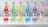 Six colorful cocktail bottles are arranged in a line on a marble surface. Each bottle is filled with a different brightly colored drink: red, green, yellow, blue, purple, and pink. The labels on the bottles display various fruit flavors, including strawberry, lemon, peach, blueberry, grape, and rose & lychee. The sleek design and vibrant colors convey a refreshing and inviting appeal.