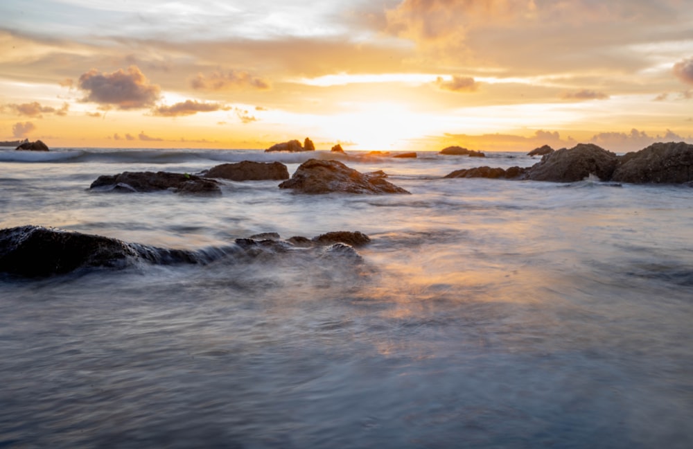 the sun is setting over the ocean with rocks in the foreground