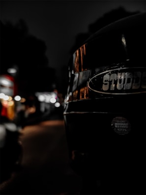 A close-up of a black helmet with the brand name 'STUDDS' prominently displayed. The background is a city street at night, blurred with bokeh lights, suggesting an urban environment. The lighting is dim, focusing on the reflective surface of the helmet.