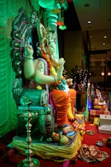 A detailed sculpture of a deity, likely Ganesha, is adorned with colorful garlands and placed on an intricately designed throne. The setting is festive, with bright decorations including flowers and fruits placed around. The atmosphere is lively and ceremonial, with an ornate lamp and red carpet adding to the celebration. The background includes draped fabrics and soft lighting.