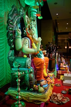 A detailed sculpture of a deity, likely Ganesha, is adorned with colorful garlands and placed on an intricately designed throne. The setting is festive, with bright decorations including flowers and fruits placed around. The atmosphere is lively and ceremonial, with an ornate lamp and red carpet adding to the celebration. The background includes draped fabrics and soft lighting.