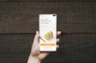 A hand holding a smartphone in front of a wooden background. The phone screen displays a financial investment application with the text 'The smartest thing to do with your money'. Below it, there are illustrations of stacked coins and a call-to-action button labeled 'Start investing'.