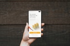 A hand holding a smartphone in front of a wooden background. The phone screen displays a financial investment application with the text 'The smartest thing to do with your money'. Below it, there are illustrations of stacked coins and a call-to-action button labeled 'Start investing'.