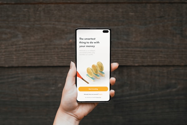 A hand holding a smartphone in front of a wooden background. The phone screen displays a financial investment application with the text 'The smartest thing to do with your money'. Below it, there are illustrations of stacked coins and a call-to-action button labeled 'Start investing'.