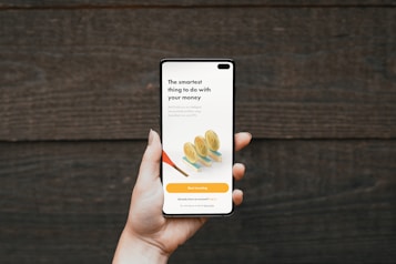 A hand holding a smartphone in front of a wooden background. The phone screen displays a financial investment application with the text 'The smartest thing to do with your money'. Below it, there are illustrations of stacked coins and a call-to-action button labeled 'Start investing'.