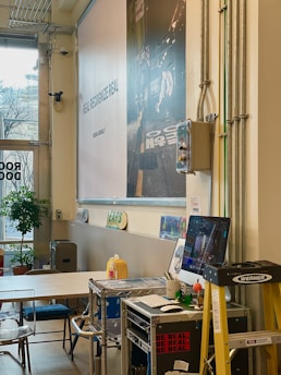 A modern workspace with a large wall graphic featuring text 'REAL RECOGNIZE REAL' in a creative font. In the foreground, there's a computer on a metal cart surrounded by various office items, including a keyboard, mouse, and a mug. A ladder is positioned nearby, and several skateboards are mounted on the wall as decoration. A potted plant is set by the large window, which allows natural light to brighten the room.