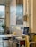 A modern workspace with a large wall graphic featuring text 'REAL RECOGNIZE REAL' in a creative font. In the foreground, there's a computer on a metal cart surrounded by various office items, including a keyboard, mouse, and a mug. A ladder is positioned nearby, and several skateboards are mounted on the wall as decoration. A potted plant is set by the large window, which allows natural light to brighten the room.