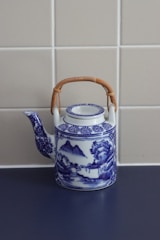 A vintage porcelain teapot with intricate blue and white designs, featuring a landscape scene with mountains and trees. It has a braided bamboo handle and sits on a dark surface against a tiled beige wall.