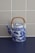 A vintage porcelain teapot with intricate blue and white designs, featuring a landscape scene with mountains and trees. It has a braided bamboo handle and sits on a dark surface against a tiled beige wall.