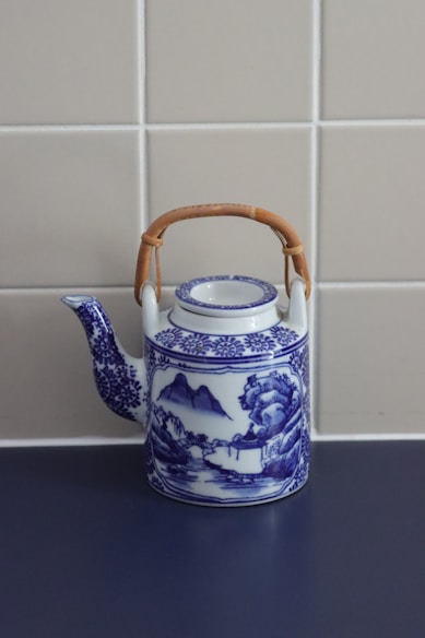 A vintage porcelain teapot with intricate blue and white designs, featuring a landscape scene with mountains and trees. It has a braided bamboo handle and sits on a dark surface against a tiled beige wall.