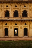 A historical building facade with intricate arched windows and doorways, displaying a blend of decorative stonework and architectural design. The structure appears aged, with a weathered and rustic texture on the walls. Light and shadows play across the surfaces, highlighting the detailed craftsmanship.