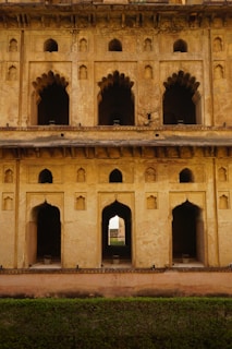 A historical building facade with intricate arched windows and doorways, displaying a blend of decorative stonework and architectural design. The structure appears aged, with a weathered and rustic texture on the walls. Light and shadows play across the surfaces, highlighting the detailed craftsmanship.