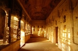 An ancient architectural space features intricately carved walls and ceilings. Sunlight streams through detailed latticework, casting patterned shadows on the floor and walls. The interior is beautifully weathered, with an earthy color palette dominated by browns and golds.